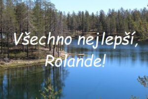 Nápis "Všechno nejlepší, Rolande!" na pozadí řeky lemované stromy jehličnatého lesa.